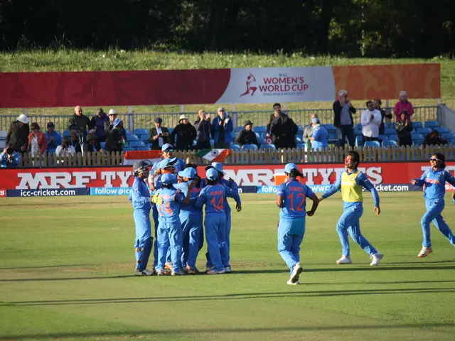 The image shows a group of women's cricket players celebrating a wicket on a grassy field. In the...