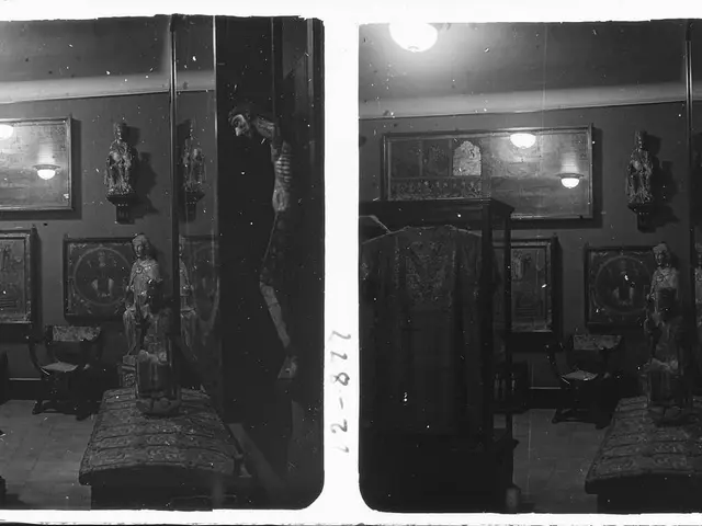 The image shows a black and white photo of a room with a bed, a chair, a table, a cupboard, a...