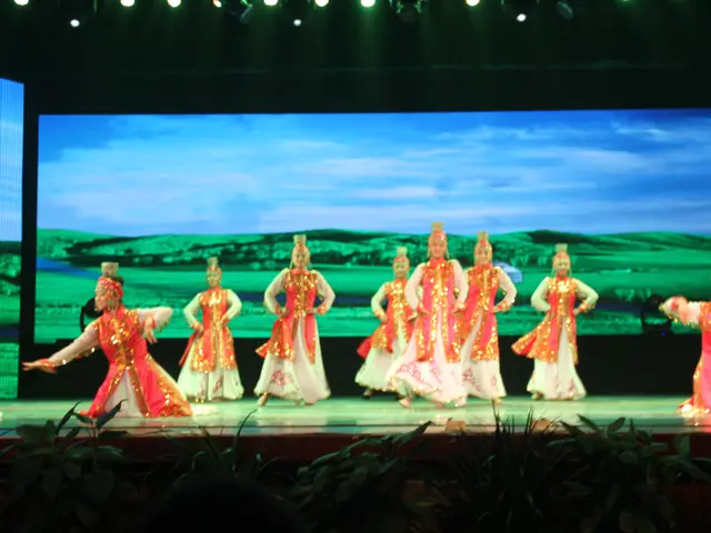 The image shows a group of people wearing colorful costumes performing on a stage, illuminated by...