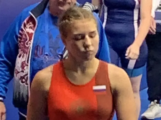 The image shows a woman in a red wrestling suit walking on a blue mat, surrounded by a group of...