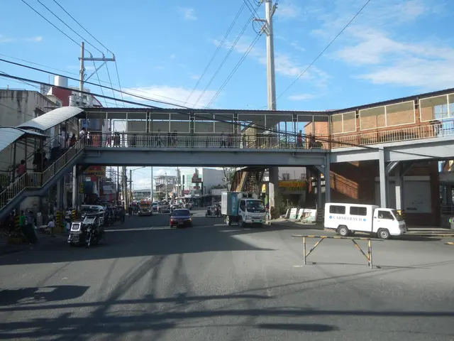 The image shows a city street with cars and trucks driving down it, a pedestrian bridge with...