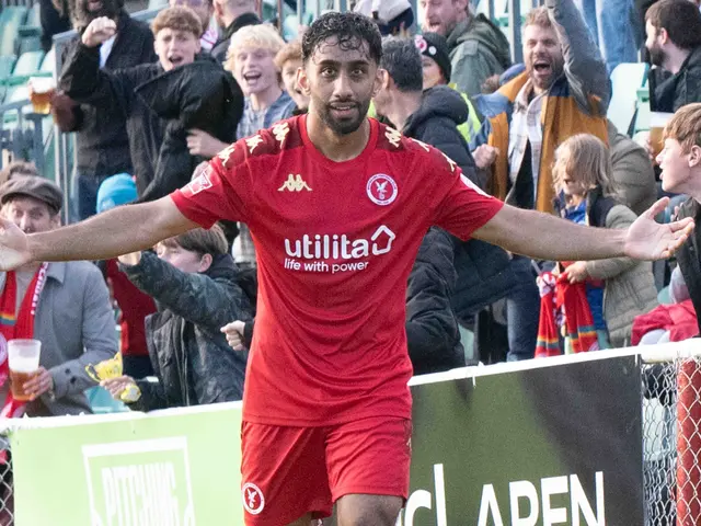 The image shows a soccer player in a red uniform running on a field with his arms outstretched,...