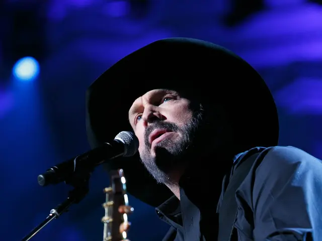 The image shows a man wearing a black shirt and a black hat, playing a guitar and singing into a...