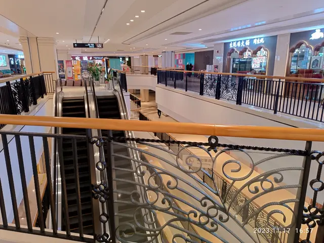 The image shows the interior of a shopping mall with escalators, railings, houseplants, stores,...