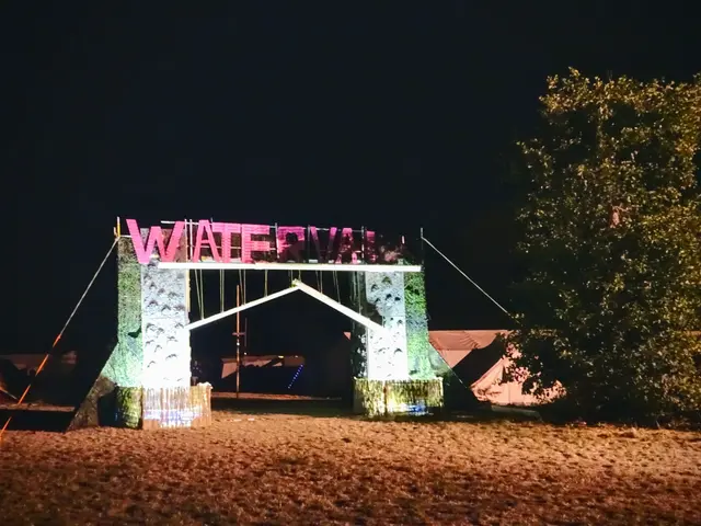 The image shows a stage set up for a music festival at night, with an arch with text on it, a tree...