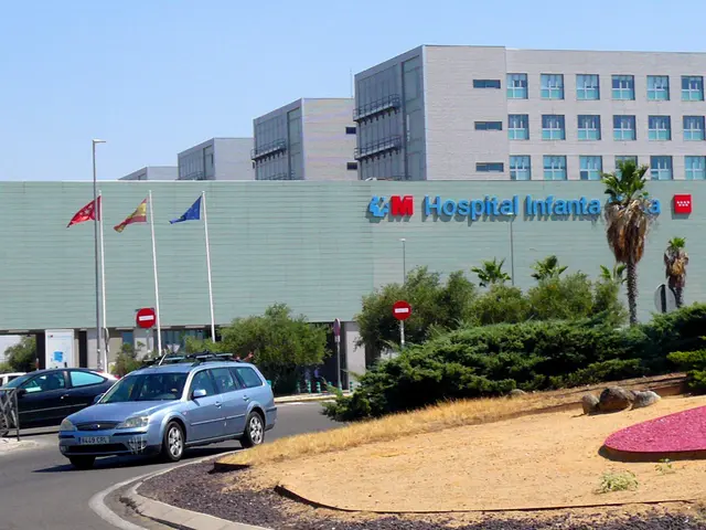The image shows a hospital with cars parked in front of it, surrounded by buildings, street poles,...