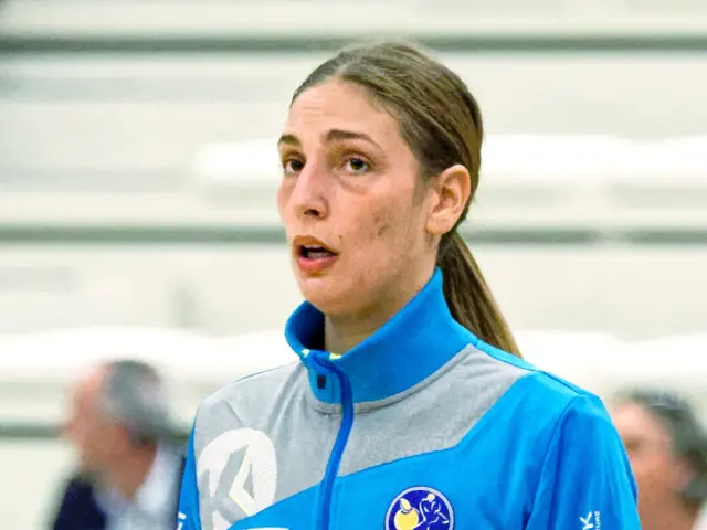 The image shows a woman in a blue jacket and purple pants standing on a basketball court, with a...