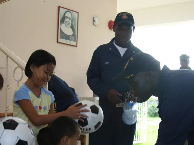 The image shows a group of children standing around a man holding a soccer ball. The man is wearing...