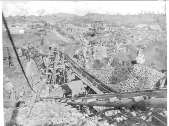 The image shows a black and white photo of a coal mine in Columbia, with a few buildings, trees,...