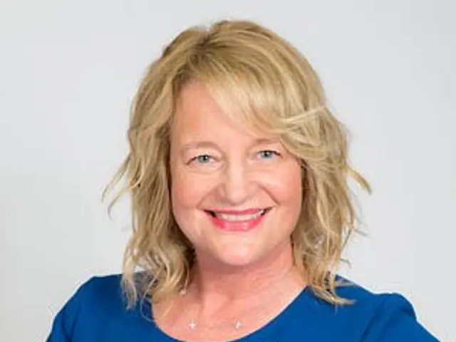The image shows a woman in a blue dress smiling at the camera against a white background. She is...
