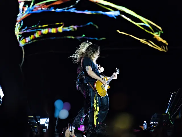 The image shows a man and a woman playing guitars on stage at a concert. The man is wearing a black...