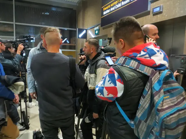 The image shows a group of people standing in an airport, some of them holding cameras and mobile...