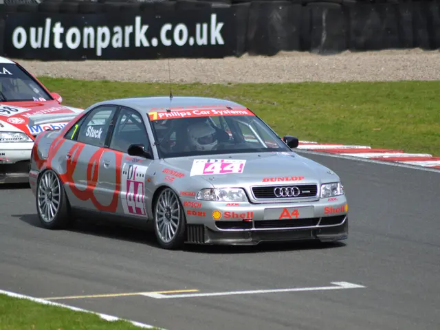 The image shows two cars racing on a race track with people wearing helmets in the background. The...
