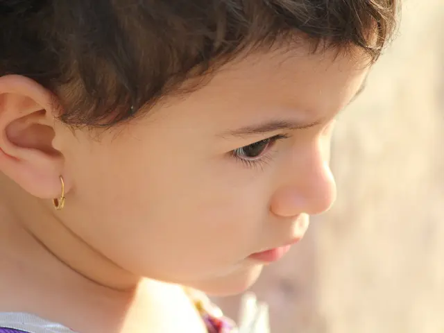 The image shows a close up of a little girl wearing a purple and white dress and gold earrings. The...