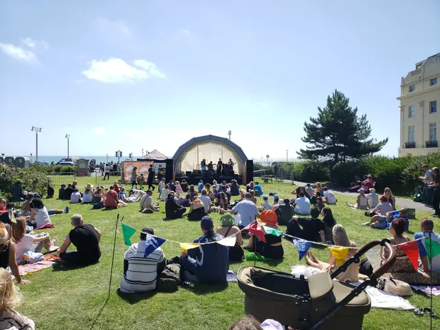 The image shows a large group of people sitting on top of a lush green field, with a stage in the...