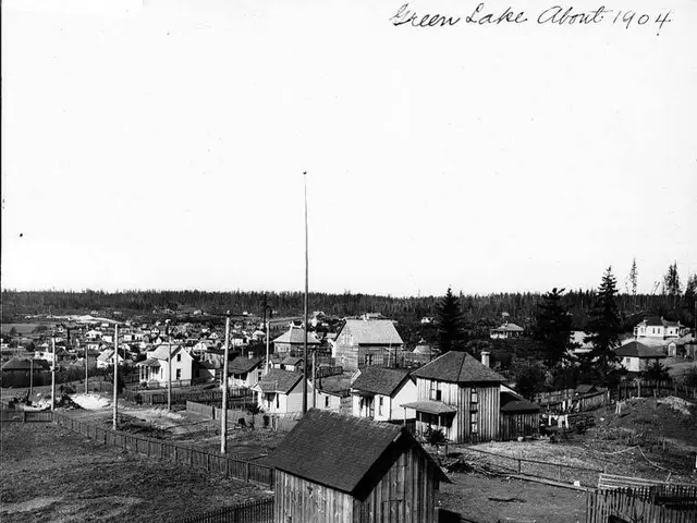 The image shows a black and white photo of a small town with houses, poles, trees, and a fence in...