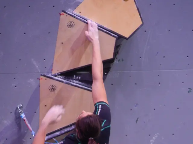 The image shows a woman climbing on a rock wall in a gym. She is wearing a harness and is using a...