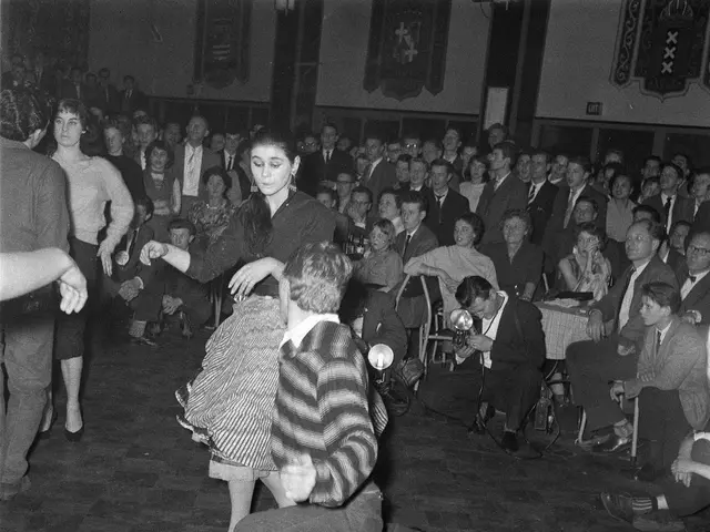The image shows a couple of people dancing in front of a crowd of people sitting on chairs and some...