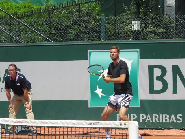 The image shows two men playing tennis on a clay court. The man in the foreground is wearing a...