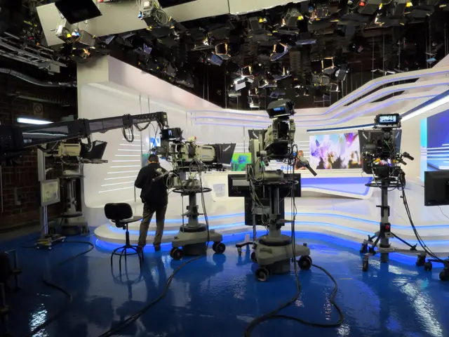 The image shows a man standing in front of a TV set with cameras on the floor, a chair, a display...