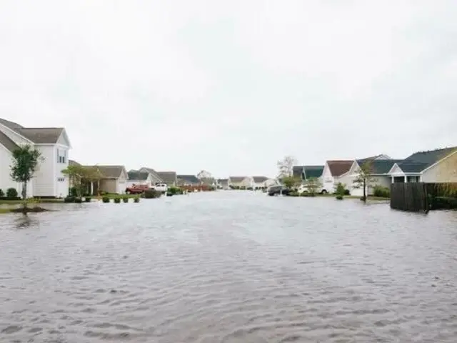 The image shows a flooded street in a residential area with cars parked on the side of it,...