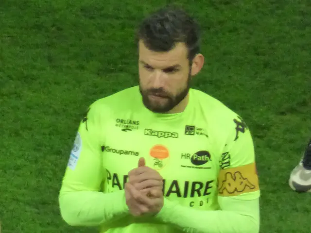 The image shows a man in a yellow goalkeeper's uniform standing on a soccer field, clapping his...