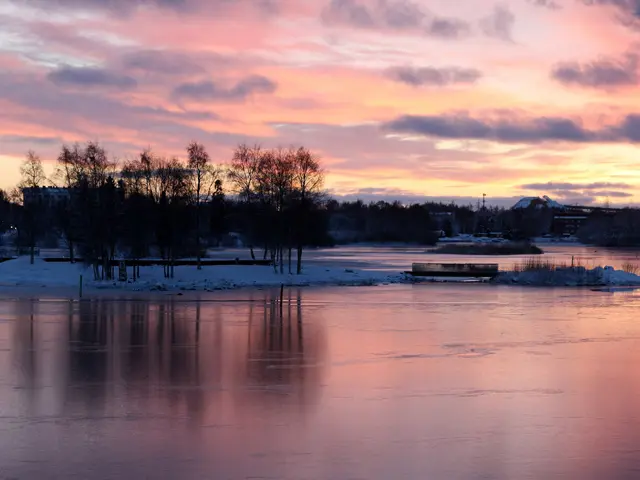 The image shows a beautiful sunset over a body of water with trees in the foreground, snow on the...
