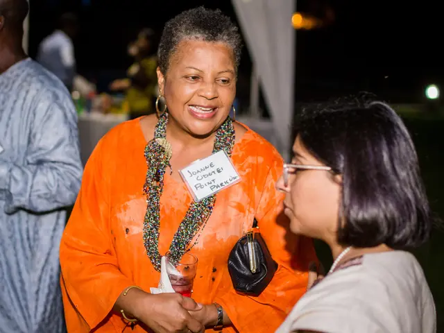 The image shows two women talking to each other at a party. The woman in the middle is wearing an...