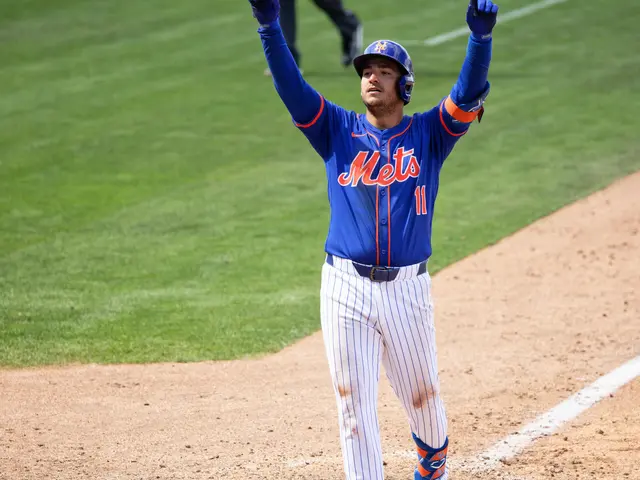 The image shows a baseball player standing on top of a field, wearing a blue t-shirt, white pants,...