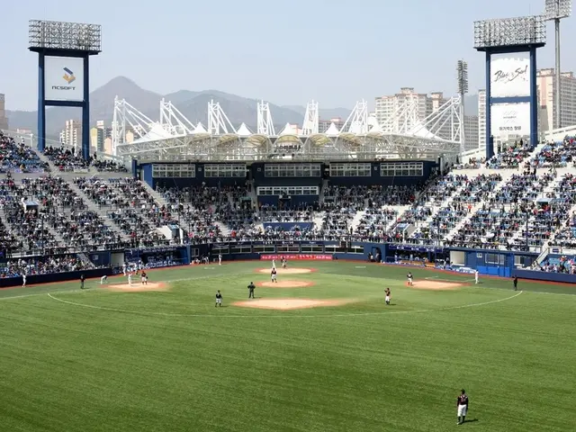The image shows a baseball game being played in a stadium, with a group of people on the ground and...