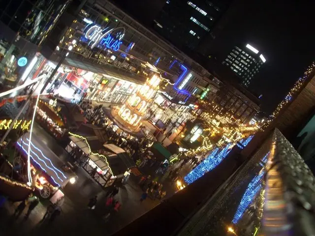 The image shows a bustling city street filled with lots of people walking around at night,...