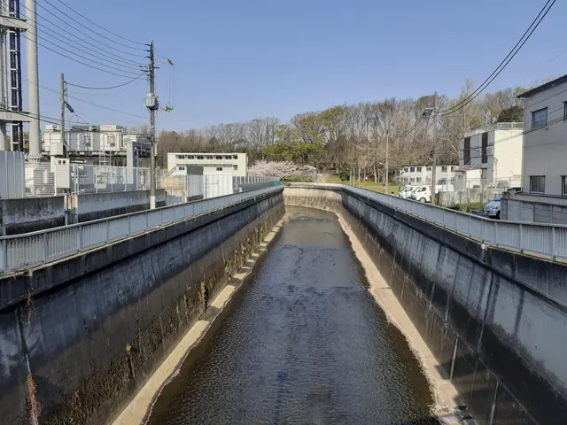 The image shows a small canal running through a city with buildings on either side, electric poles...