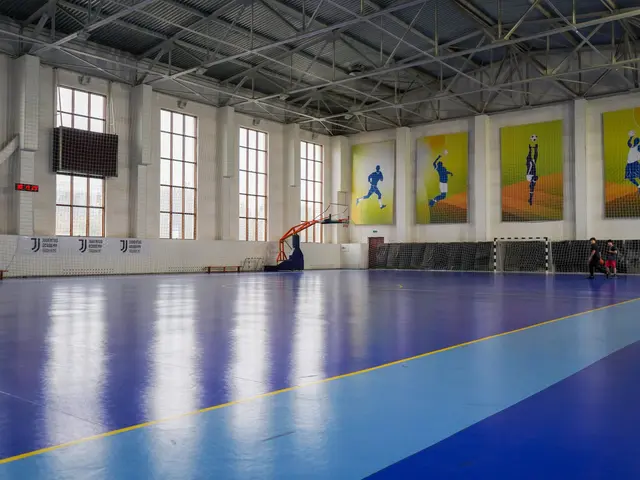 The image shows a large indoor futsal court with a blue and yellow floor, a goal post, a net, a...