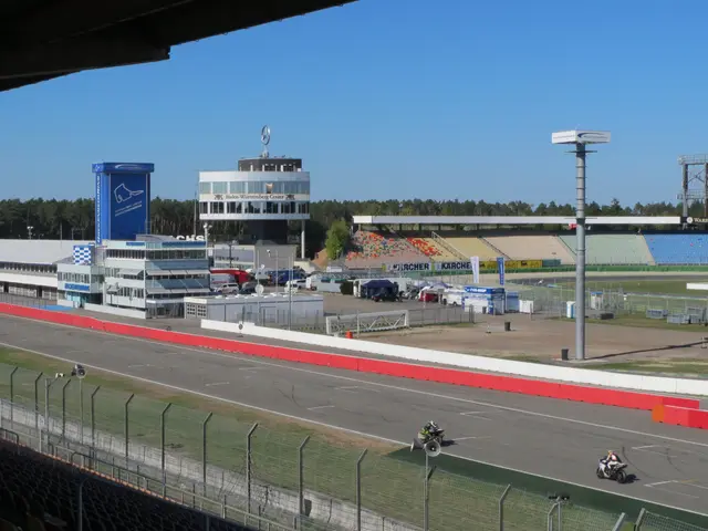 The image shows a group of people riding motorcycles on top of a race track, surrounded by a metal...