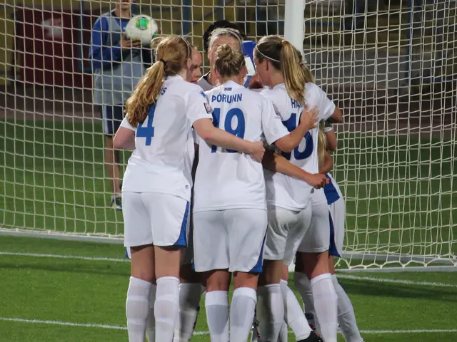 The image shows a group of women's soccer players huddled together on the field, wearing sports...