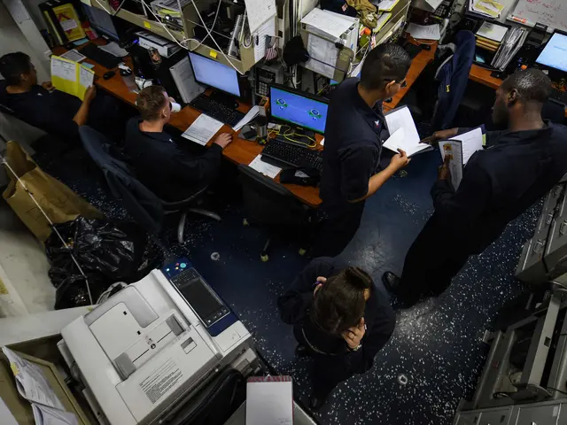 The image shows a group of people sitting at desks in an office, surrounded by computers,...
