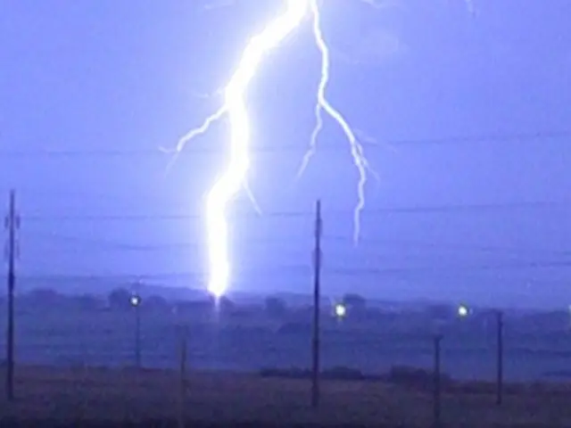 The image shows a lightning storm over a field with power lines in the background, illuminated by...