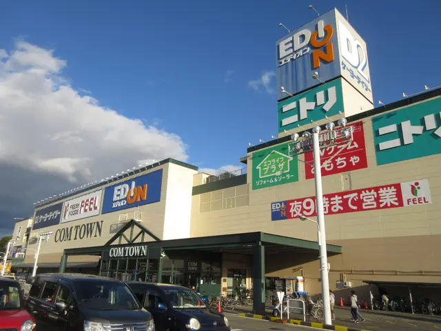 The image shows a shopping center with cars parked in front of it, surrounded by buildings, name...