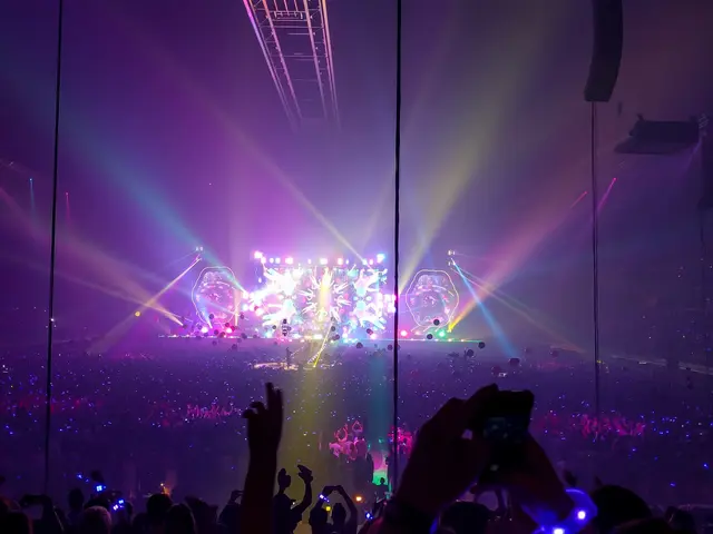 The image shows a crowd of people at a concert with their hands in the air, illuminated by colorful...