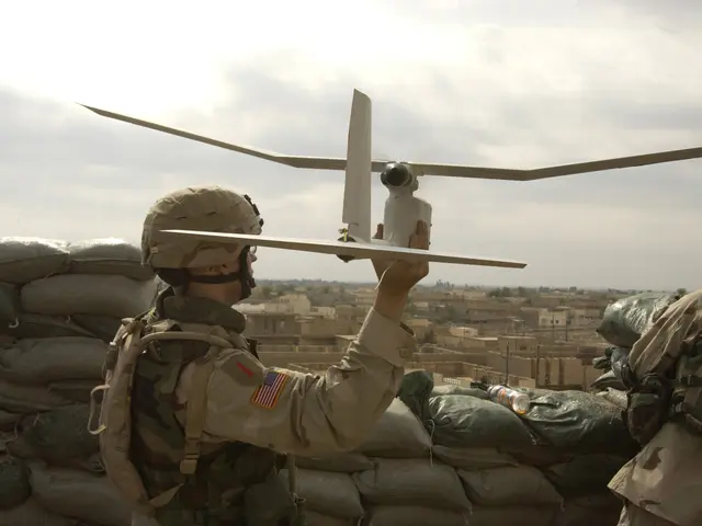 The image shows two soldiers wearing helmets and bags, holding up a drone in front of a pile of...