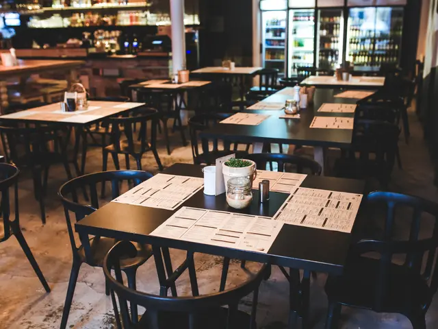 The image shows a restaurant with tables and chairs in front of a bar, with menu cards, glasses,...