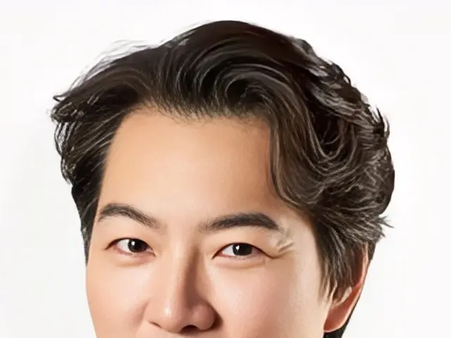 The image shows a man in a blue shirt smiling at the camera against a white background. He appears...