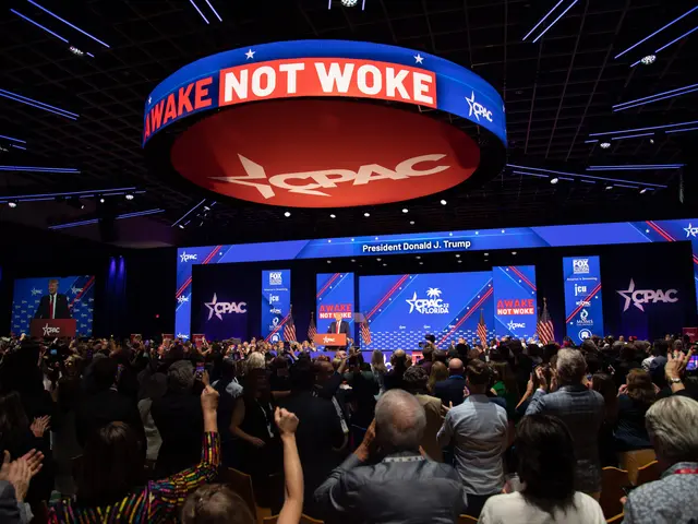 The image shows a large crowd of people sitting in front of a stage with a sign that reads "Wake...