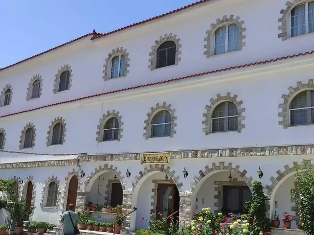 The image shows a large white building with many windows and a flag on top of it, surrounded by...