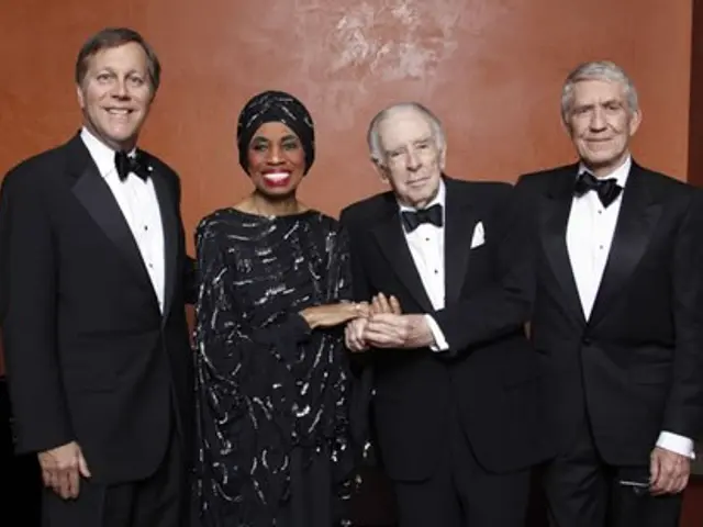 The image shows a group of four people in tuxedos standing next to each other, smiling and posing...