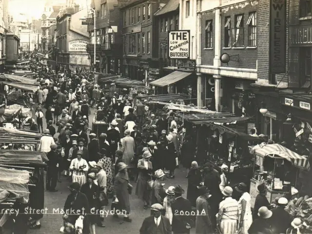 The image shows a black and white photo of a bustling street market in Croydon, with a number of...