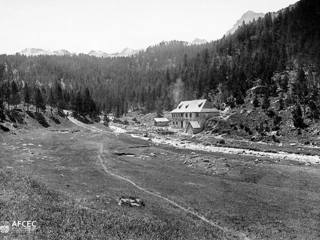 The image shows a black and white photo of a small village nestled in the mountains, with a house...