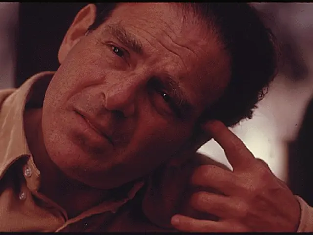 The image shows a man with his hand on his chin, looking directly at the camera with a blurred...