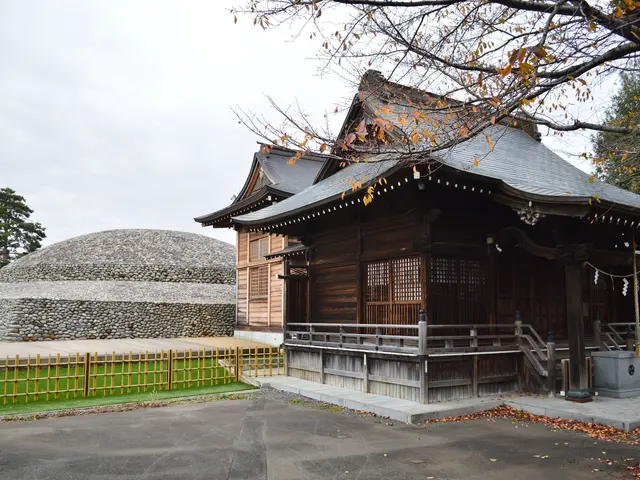The image shows a large wooden building with a tree in front of it, surrounded by a fence, grass,...
