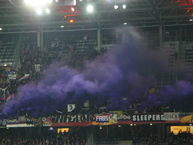The image shows a large crowd of people in a stadium with purple smoke billowing out of them. There...